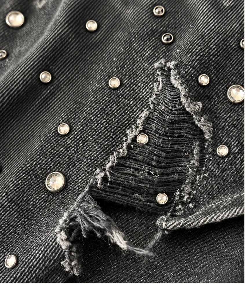 Close-up of black denim with controlled distressing, frayed raw edges and scattered metal embellishment.